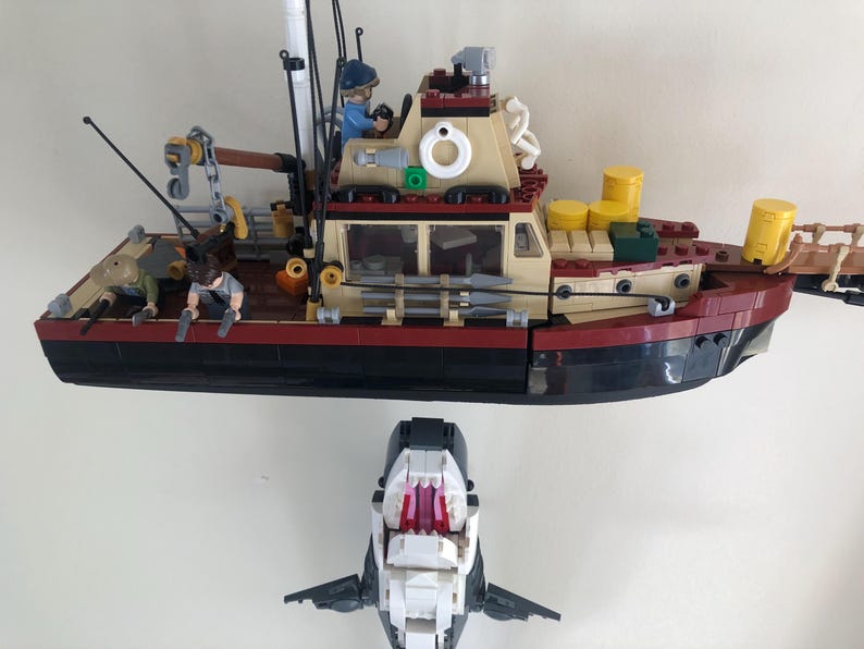 May include: A Lego model of a fishing boat with a red, black, and tan hull, and a shark model below. The boat has figures on deck, including one in the cockpit. The shark has an open mouth with red teeth.