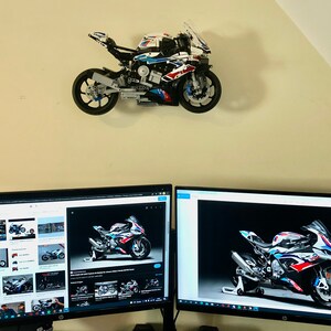 May include: A white, blue, and red Lego motorcycle model mounted on a wall, with two computer monitors displaying images of the same motorcycle.