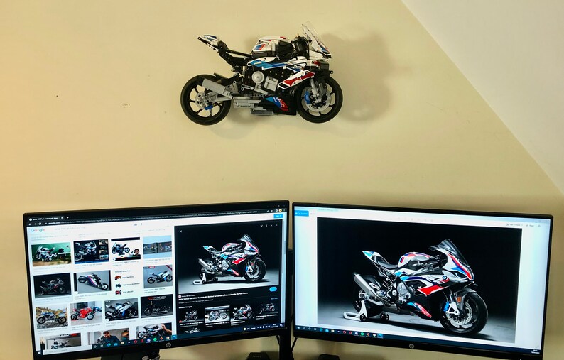 May include: A white, blue, and red Lego motorcycle model mounted on a wall, with two computer monitors displaying images of the same motorcycle.