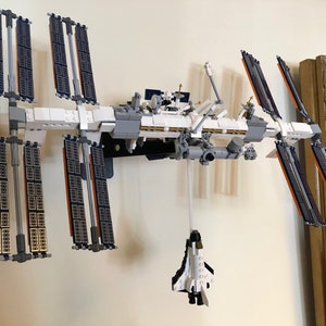 May include: A white and gray Lego model of the International Space Station with solar panels and a space shuttle attached.