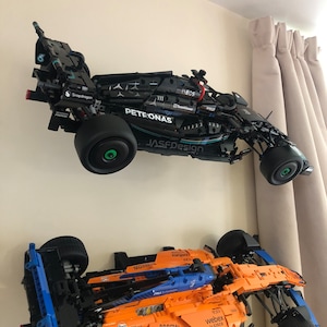 May include: Two Lego Formula 1 race cars mounted on a wall. The top car is black with blue and silver accents and the text "PETRONAS" and "JASFDesign". The bottom car is orange with blue accents and the text "webex by CISCO" and "MOWN".