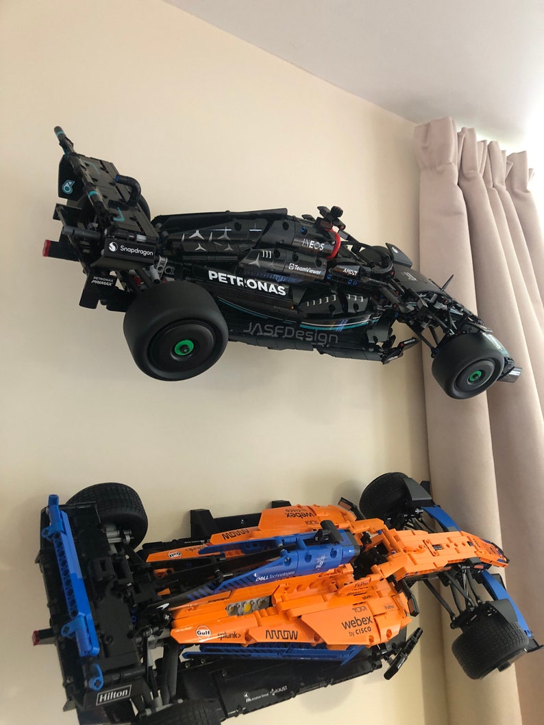 May include: Two Lego Formula 1 race cars mounted on a wall. The top car is black with blue and silver accents and the text "PETRONAS" and "JASFDesign". The bottom car is orange with blue accents and the text "webex by CISCO" and "MOWN".