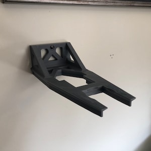 May include: A black plastic wall mount for a video game console. The mount is shaped like a letter H and has a rectangular opening in the center.