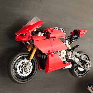 Op de afbeelding: Een rood en zwart Lego-motorfietsmodel met een heldere voorruit. De motorfiets heeft een gedetailleerde motor en een kettingaandrijving.