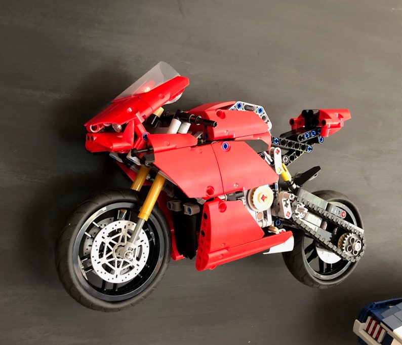 Op de afbeelding: Een rood en zwart Lego-motorfietsmodel met een heldere voorruit. De motorfiets heeft een gedetailleerde motor en een kettingaandrijving.