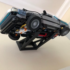 May include: A black plastic stand holds a gray and silver Lego DeLorean time machine model, suspended in mid-air, with the underside of the car facing the viewer.