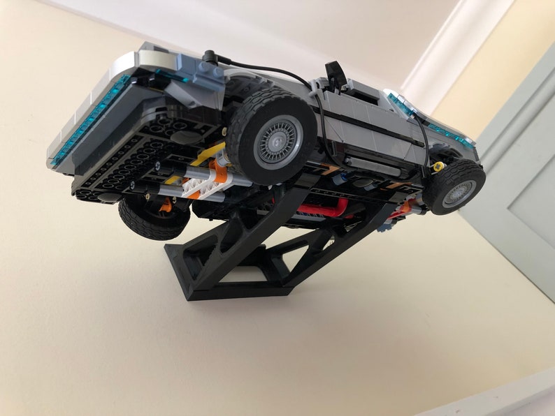 May include: A black plastic stand holds a gray and silver Lego DeLorean time machine model, suspended in mid-air, with the underside of the car facing the viewer.