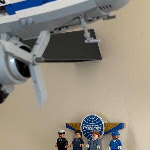 May include: A LEGO model of a vintage airplane with white and blue coloring, mounted on a wall. Below, a LEGO display features four miniature figures in uniforms, with the Pan Am logo prominently displayed.