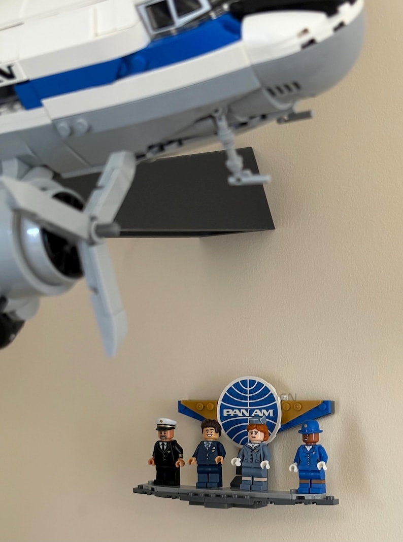 May include: A LEGO model of a vintage airplane with white and blue coloring, mounted on a wall. Below, a LEGO display features four miniature figures in uniforms, with the Pan Am logo prominently displayed.