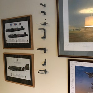 May include: A white wall with six gray metal wall mounted hooks. The hooks are in a vertical line. There are three framed pictures on the wall. The pictures are of airplanes.