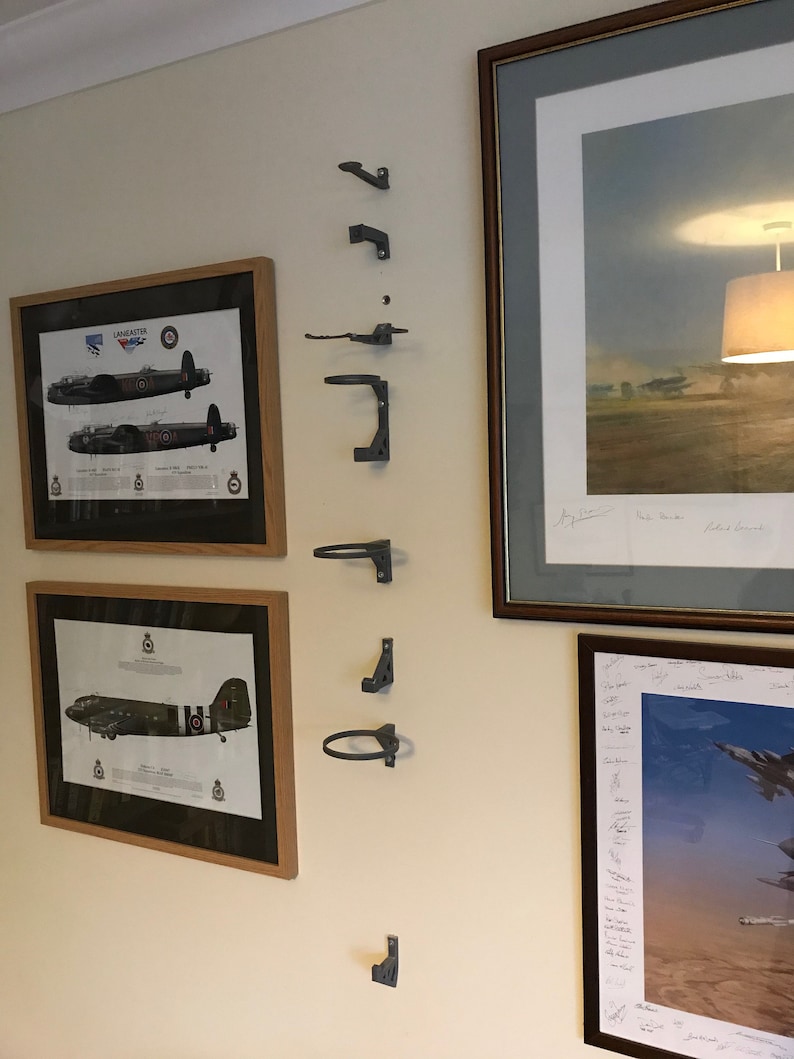 May include: A white wall with six gray metal wall mounted hooks. The hooks are in a vertical line. There are three framed pictures on the wall. The pictures are of airplanes.