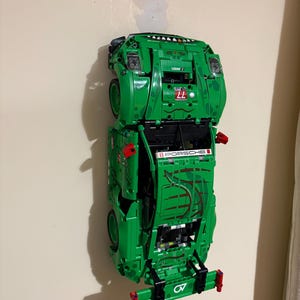 May include: A green Lego Porsche race car model mounted on a wall. The model features the number 77, the word "PORSCHE", and other racing decals. The car is primarily green with red and black accents.