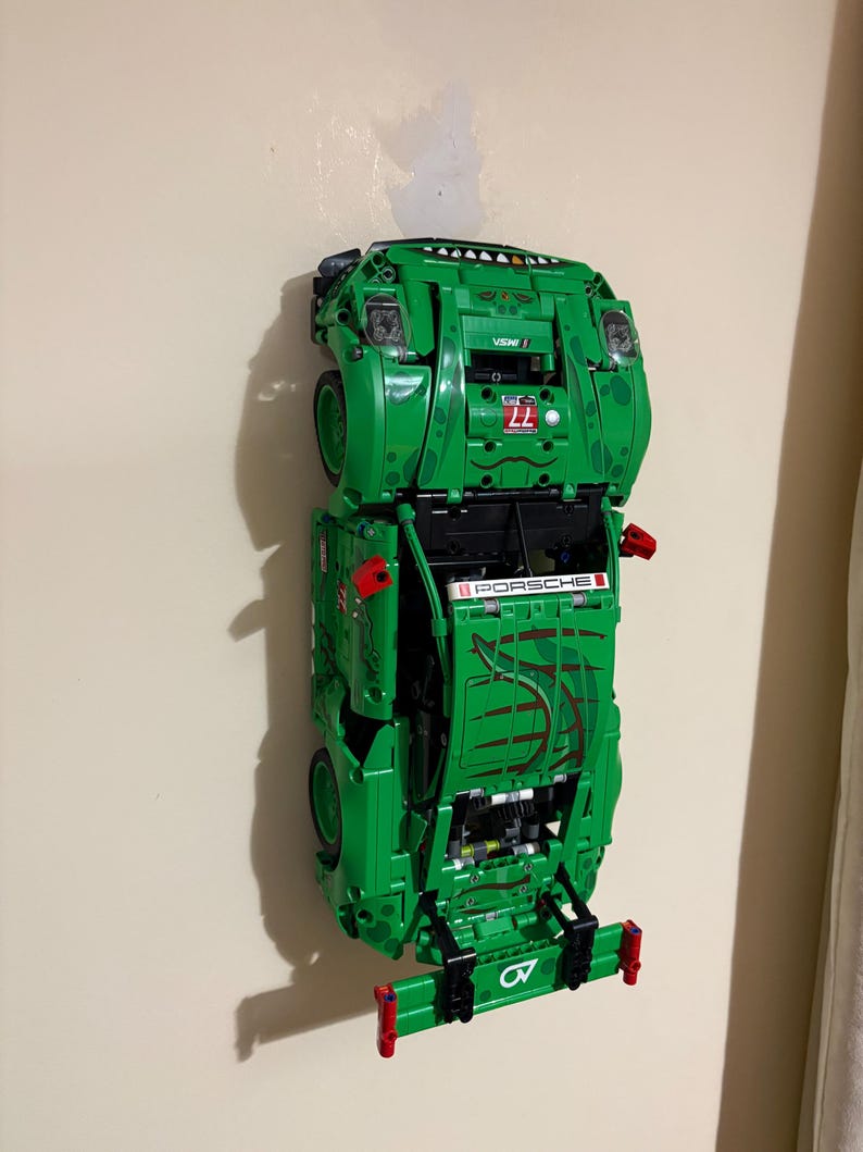 May include: A green Lego Porsche race car model mounted on a wall. The model features the number 77, the word "PORSCHE", and other racing decals. The car is primarily green with red and black accents.