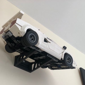 May include: A white Lego Lamborghini Countach model car displayed on a black wall mount. The car is suspended in mid-air, as if it is driving up the wall. The wall mount is labeled "MODERN COUNTACH".