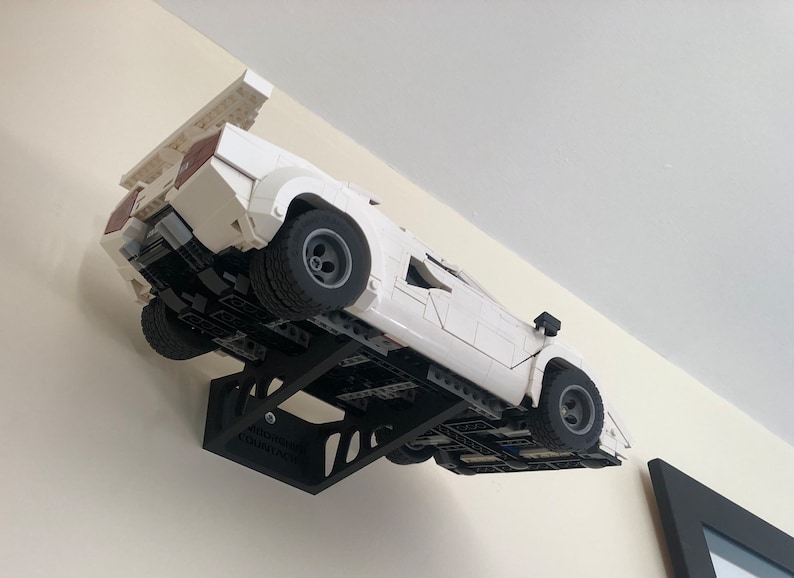 May include: A white Lego Lamborghini Countach model car displayed on a black wall mount. The car is suspended in mid-air, as if it is driving up the wall. The wall mount is labeled "MODERN COUNTACH".