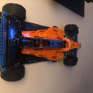 Puede incluir: Un modelo de coche de carreras de F&oacute;rmula 1 McLaren Lego negro y naranja con las palabras "WEBEX" y "M2000" impresas en el lateral. El coche est&aacute; montado en una pared.