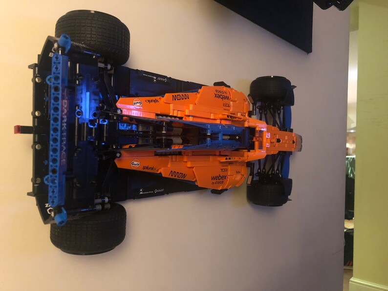 Puede incluir: Un modelo de coche de carreras de F&oacute;rmula 1 McLaren Lego negro y naranja con las palabras "WEBEX" y "M2000" impresas en el lateral. El coche est&aacute; montado en una pared.