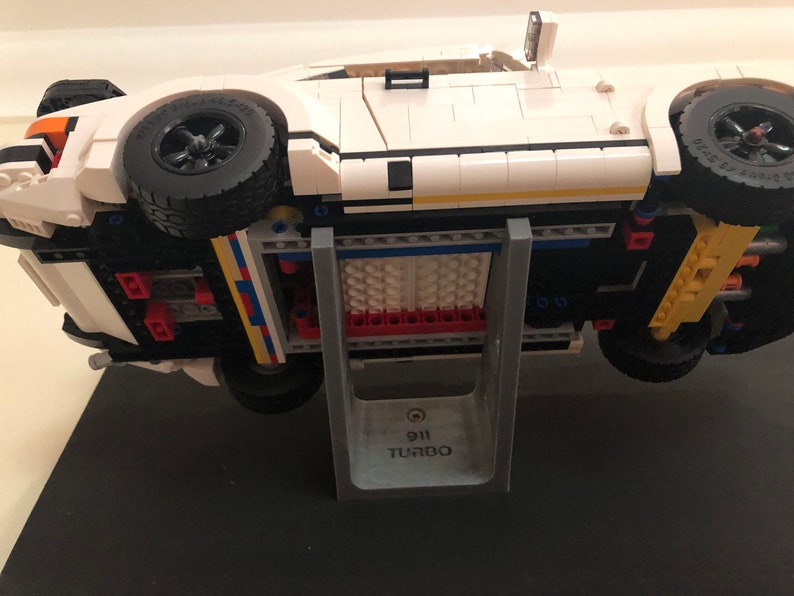 May include: A white Lego Porsche 911 Turbo model car on a gray stand with the text "911 Turbo" printed on the stand.
