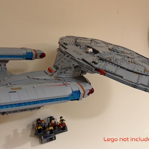 Puede incluir: Maqueta de nave espacial gris con detalles azules y rojos, montada en la pared. El modelo va acompa&ntilde;ado de un peque&ntilde;o grupo de figuras. Tambi&eacute;n se ve un reloj redondo con marco negro. El texto "Lego not included" est&aacute; en la parte inferior.