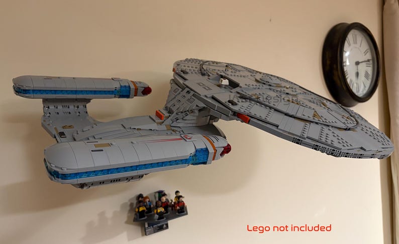Puede incluir: Maqueta de nave espacial gris con detalles azules y rojos, montada en la pared. El modelo va acompa&ntilde;ado de un peque&ntilde;o grupo de figuras. Tambi&eacute;n se ve un reloj redondo con marco negro. El texto "Lego not included" est&aacute; en la parte inferior.