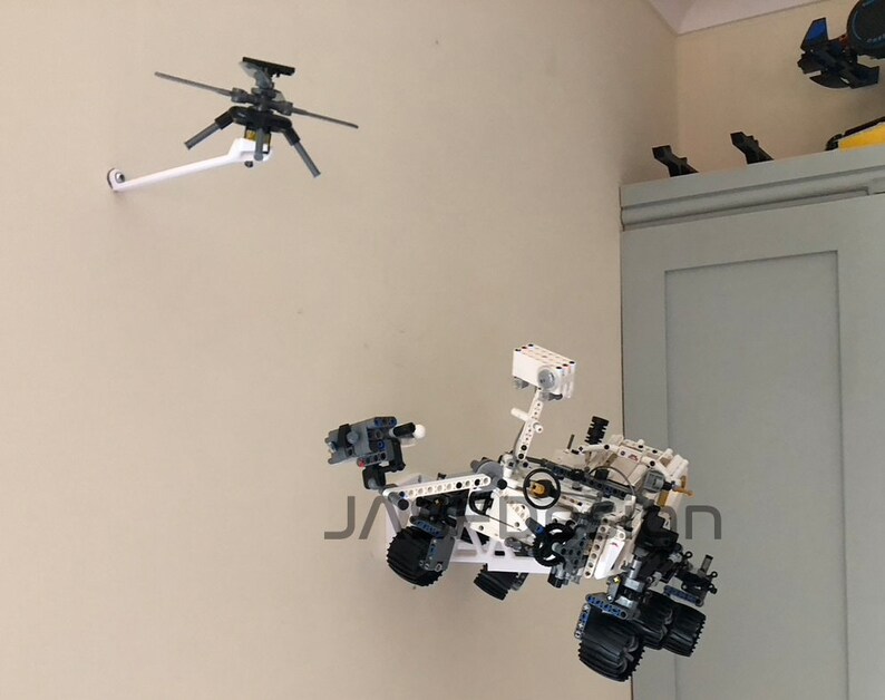 May include: A white Lego model of the Mars rover Perseverance, with a white Lego helicopter hovering above it. Both models are mounted on a white wall.