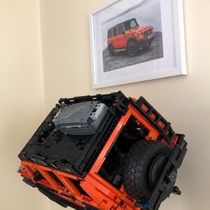 May include: An orange and black Lego model of a Mercedes-Benz G-Wagon, with a spare tire mounted on the back. The model is suspended in mid-air, as if it is floating.
