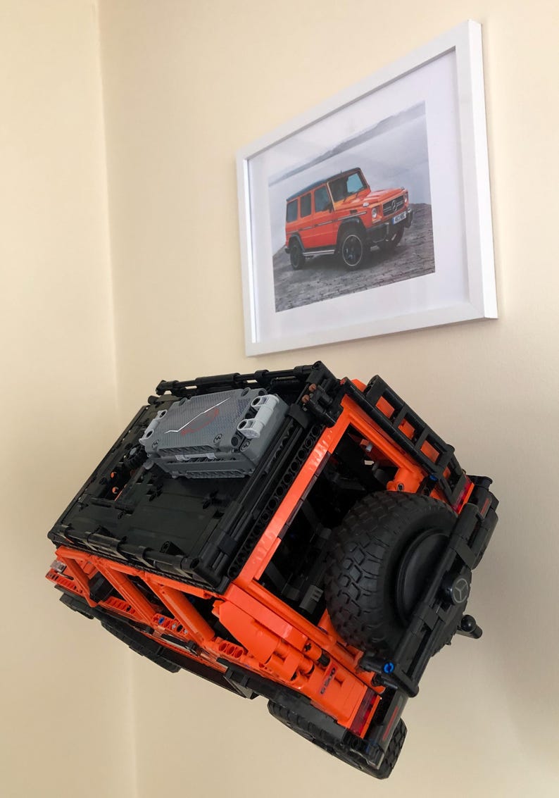 May include: An orange and black Lego model of a Mercedes-Benz G-Wagon, with a spare tire mounted on the back. The model is suspended in mid-air, as if it is floating.