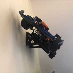 May include: A black and blue Lego race car model mounted on a black wall mount. The car has orange accents and is displayed in a tilted position.