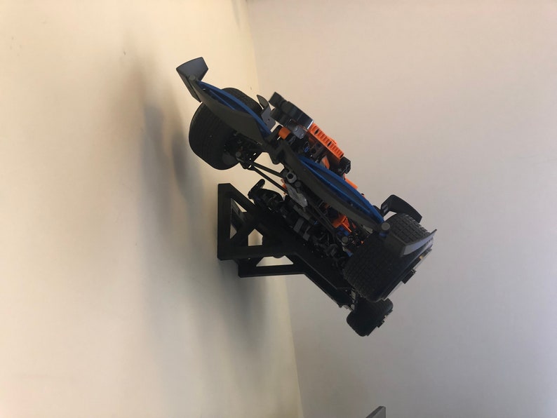 May include: A black and blue Lego race car model mounted on a black wall mount. The car has orange accents and is displayed in a tilted position.