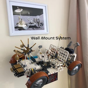 May include: A white framed picture of a lunar rover on a wall, with a Lego model of a lunar rover mounted below. The Lego model is white, brown, and gray, with black tires. The text "Wall Mount System" and "LEGO Not Included" are visible.