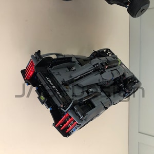May include: A black and gray Lego model of a Peugeot car with red and blue accents. The car is suspended in the air, facing the camera.