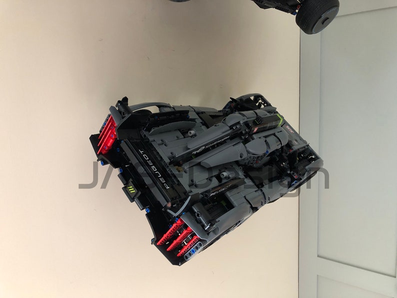 May include: A black and gray Lego model of a Peugeot car with red and blue accents. The car is suspended in the air, facing the camera.