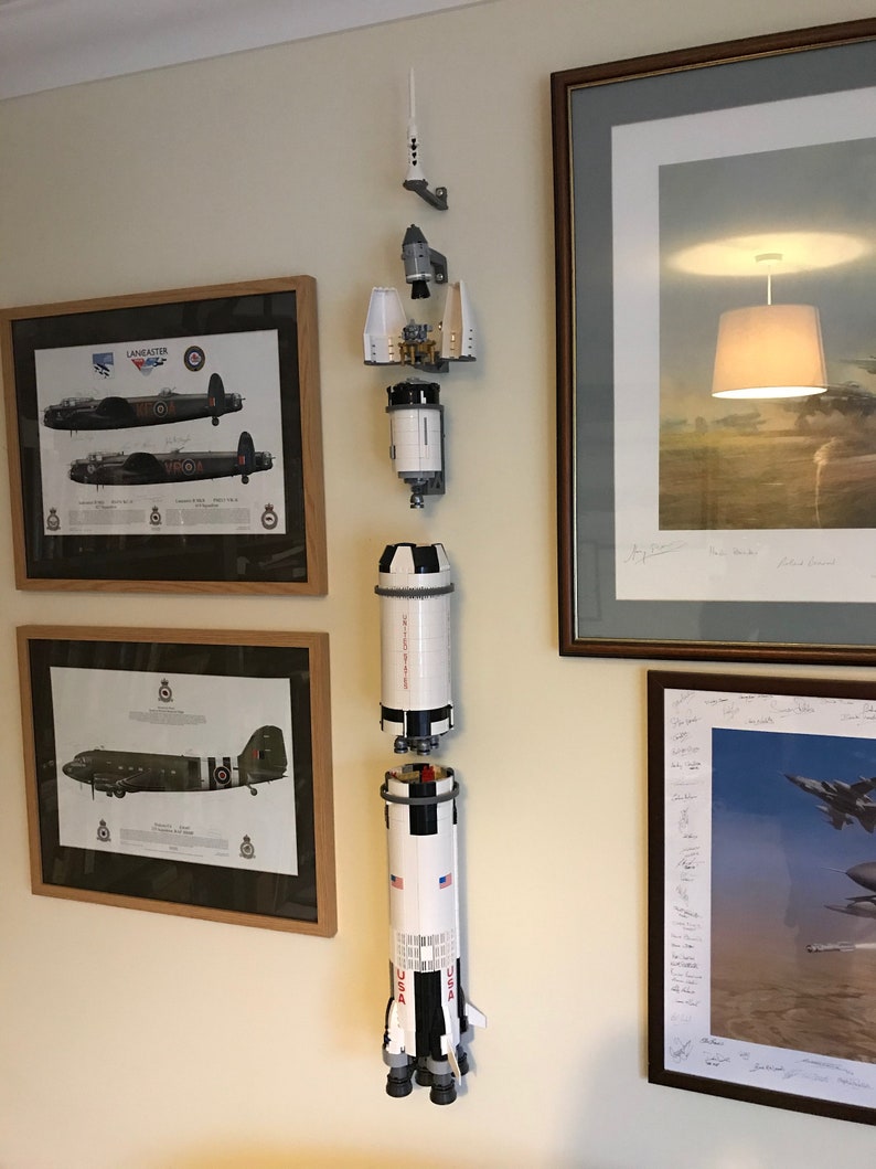 May include: A white and black Lego Saturn V rocket model is displayed on a wall, with the different sections of the rocket separated. The rocket is mounted on a white wall with framed pictures of airplanes behind it.