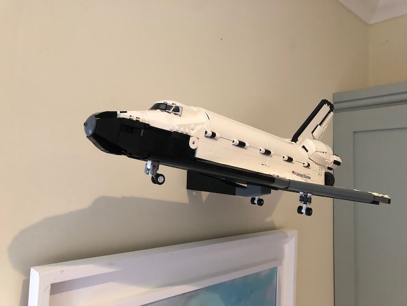 May include: A white and black model of a space shuttle, mounted on a wall. The model has the words "United States" printed on the side. The shuttle is displayed above a framed picture.