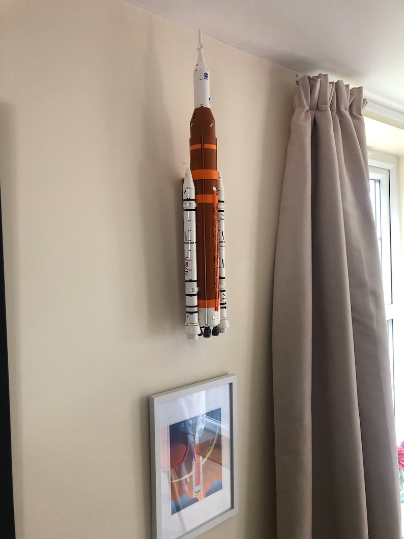 May include: A white and orange model rocket is mounted on a wall. The rocket has multiple stages and is decorated with black and white markings. The rocket is mounted on a white wall with a white picture frame below it.