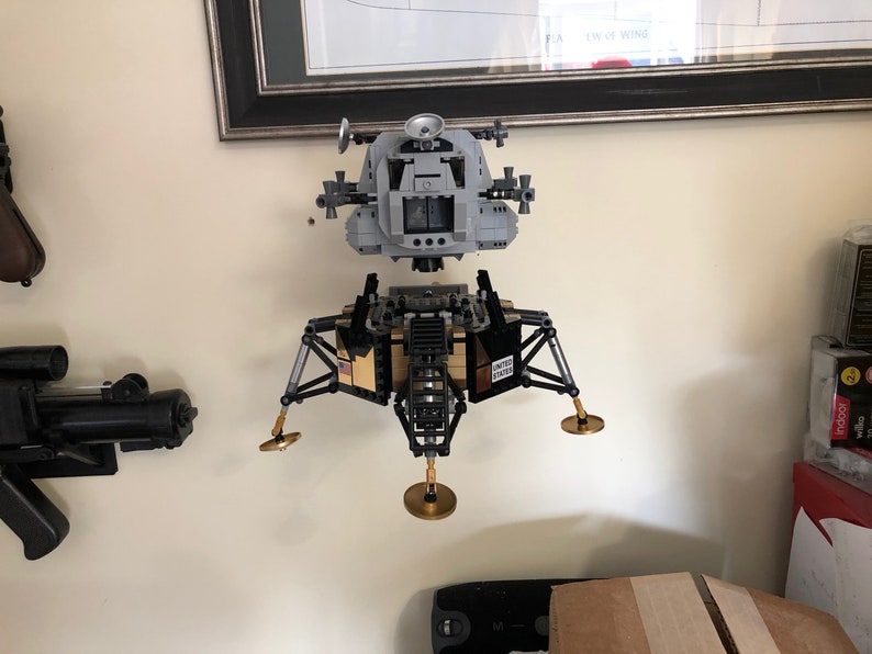 May include: A gray and black Lego model of the Apollo Lunar Module, with gold landing legs, is mounted on a wall.