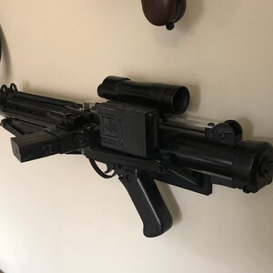 May include: A black plastic replica of a blaster weapon from the Star Wars franchise. The weapon is mounted on a wall with a white plastic mount.