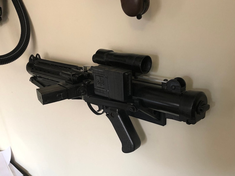 May include: A black plastic replica of a blaster weapon from the Star Wars franchise. The weapon is mounted on a wall with a white plastic mount.
