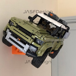 May include: A green Lego Land Rover Defender model with a white roof and black accents is mounted on a wall with an orange bracket. The model has a detailed interior and exterior, including a spare tyre on the back and a roof rack.