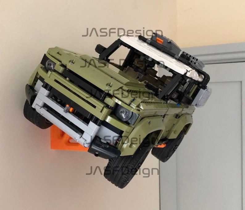 May include: A green Lego Land Rover Defender model with a white roof and black accents is mounted on a wall with an orange bracket. The model has a detailed interior and exterior, including a spare tyre on the back and a roof rack.