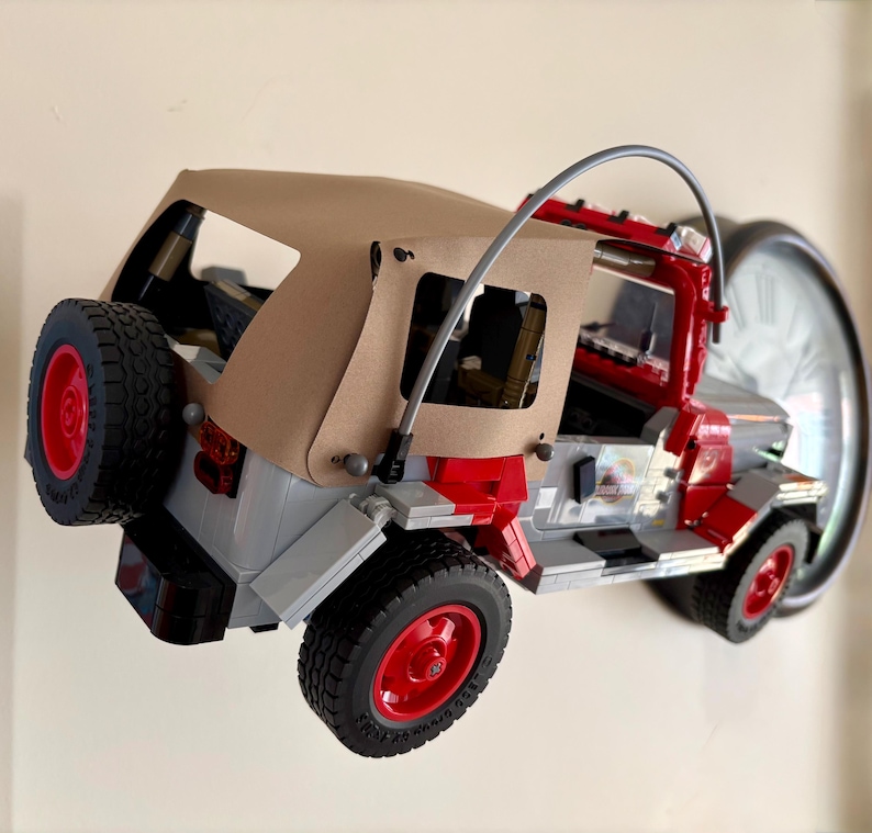 May include: A building block model of a red and gray off-road vehicle featuring a tan fabric roof, a spare tire mounted on the rear, black treaded tires with red rims, and a curved gray roll bar structure extending over the open cabin area.