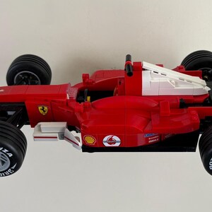 May include: A red and white Formula 1 race car model with black tires. The model features sponsor logos, including Shell, Bridgestone, and AMD. The car has a distinctive aerodynamic design.