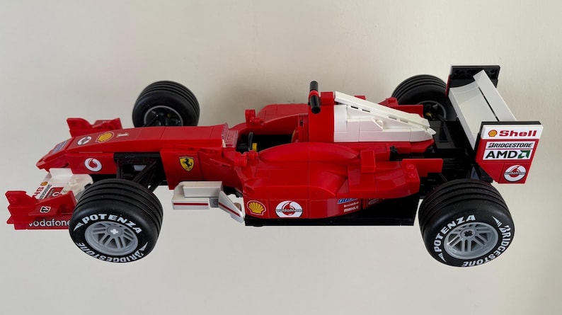 May include: A red and white Formula 1 race car model with black tires. The model features sponsor logos, including Shell, Bridgestone, and AMD. The car has a distinctive aerodynamic design.