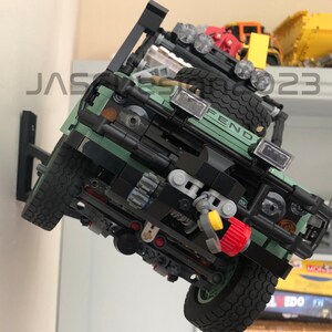May include: A green Lego Land Rover Defender model with black accents and a yellow tow hook. The model is mounted on a wall with a black bracket. The model has a FEND logo on the front.