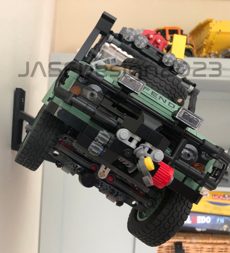 May include: A green Lego Land Rover Defender model with black accents and a yellow tow hook. The model is mounted on a wall with a black bracket. The model has a FEND logo on the front.