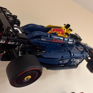 May include: A blue and black Lego model of a Formula One race car with the Red Bull logo and Mobil 1 sponsorship. The car is mounted on a wall and has a yellow and red spoiler.