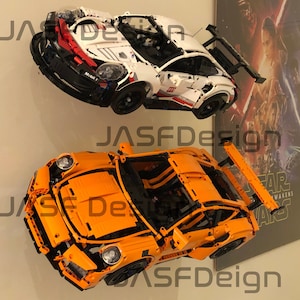 May include: Two Lego Porsche 911 GT3 RS models, one white and one orange, are displayed on a wall. The white model has a red and black stripe on the side and the text "Mobil 1" on the side. The orange model has a black rear wing.