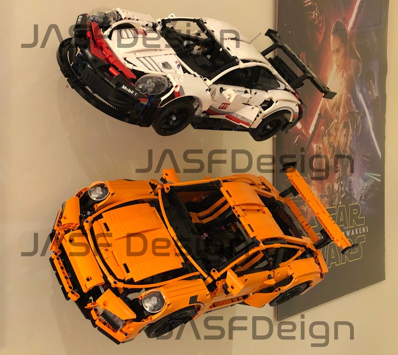 May include: Two Lego Porsche 911 GT3 RS models, one white and one orange, are displayed on a wall. The white model has a red and black stripe on the side and the text "Mobil 1" on the side. The orange model has a black rear wing.