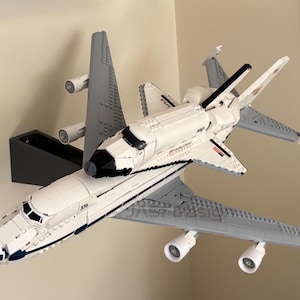 May include: A white and gray model of a space shuttle atop a gray airplane. The space shuttle has black accents and the word "USA" printed on the side. The airplane has four engines and is mounted on a black stand.