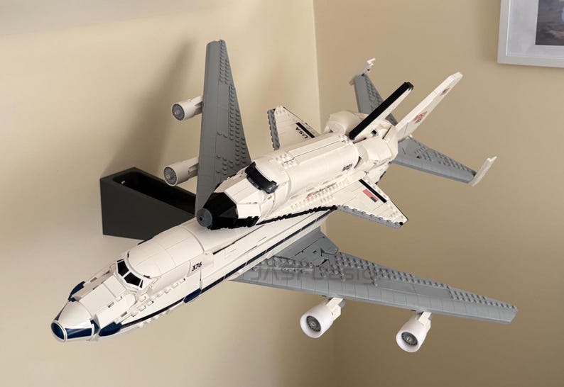 May include: A white and gray model of a space shuttle atop a gray airplane. The space shuttle has black accents and the word "USA" printed on the side. The airplane has four engines and is mounted on a black stand.